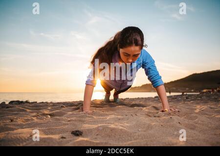 Young woman in sportswear doing push-ups on the beach. View from behind the head. In the background of the sunset and the sea. Stock Photo