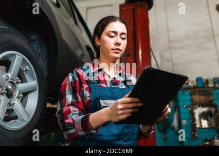 Portrait of young Caucasian female mechanic, in uniform, uses a tablet in her hands. Car on the lift in the background. Bottom view. Stock Photo