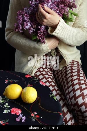 Woman holding a bouquet of blooming lilac with a hand gesture sitting at the table with lemons on it, body part crop, closeup, copy space, vertical Stock Photo