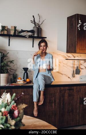 Brunette woman in pajamas eating pastries in the kitchen Stock Photo