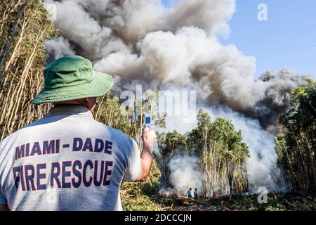 Florida,Miami-Dade Fire Rescue,Everglades,fireman using cell mobile phone camera taking photo,fire smoke burning trees controlled burn firefighter Stock Photo