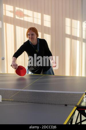 A front view of a woman with red hair wearing a black cardigan over a blue denim dress playing table tennis at home Stock Photo