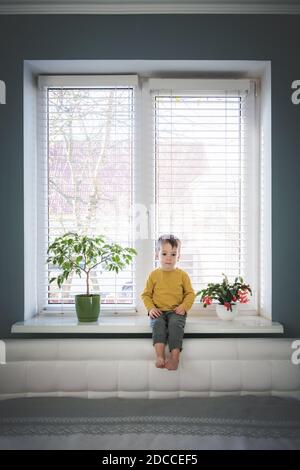 Little loneliness kid boy sitting on windowsill near large window in his room. Childhood concept Stock Photo