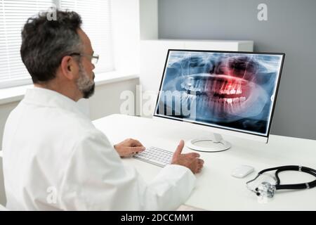Mature Male Dentist Looking At Teeth X-ray On Computer In Clinic Stock Photo