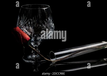 Empty crystal brandy glass & vase with a rose on a dark black background in dramatic lighting. Isolated fine dining drinking glass Stock Photo