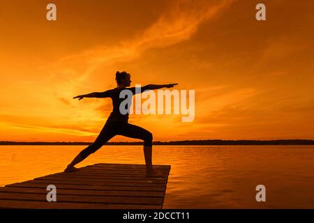 Woman practicing yoga posture Virabhadrasana II / Warrior Pose, lunging standing asana on jetty at lake at sunset