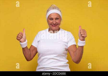 Active sporty elderly woman holding her thumb up approving your choice. Studio shot on yellow wall. Stock Photo