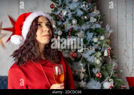 Young woman in Santa Claus hat with glass of champagne standing near Christmas tree. Charming brunette posing at coniferous tree with decorative adorn Stock Photo