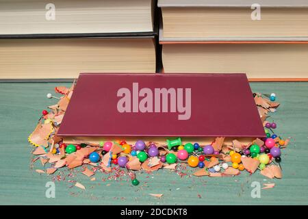 Close up shot of a small red book with colored pencil shavings and thumbtacks around it and some large books behind it. Stock Photo