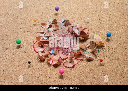 A group of pins of different sizes and colors gathered in a circle around colored pencil shavings on a cork board. Stock Photo