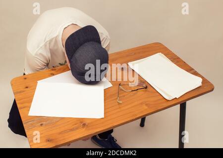 A young man sits with his head against a wooden table full of blank papers and one eyeglass. Mental health concept. Stock Photo