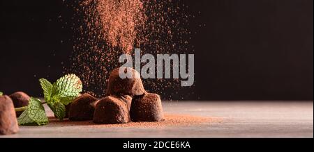 Chocolate truffles with mint leaves on wooden table and dark background falling cocoa powder. Front view. Horizontal panoramic composition. Stock Photo