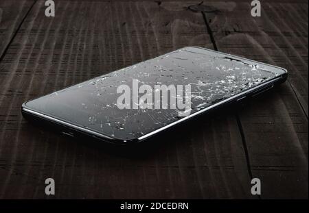 Mobile smartphone with broken touch screen on the old wooden table. LCD with many cracks and little pieces of glass. Dark tone, phone repair concept. Stock Photo