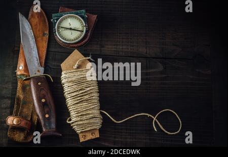 Old rusty hunting bushcraft knife, military compass and a linen rope on the wooden table. Leather cases, copy space, top view, survival concept. Stock Photo