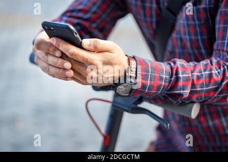 Young man wearing shirt leaning on electric scooter handlebar, holding mobile smartphone in his hands, closeup detail Stock Photo