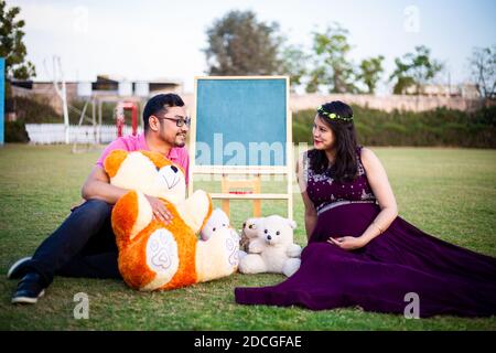 Young pregnant asian indian couple looking at each other sitting in a garden with lots teddy bear toys and blank chalkboard to write message. pregnanc Stock Photo