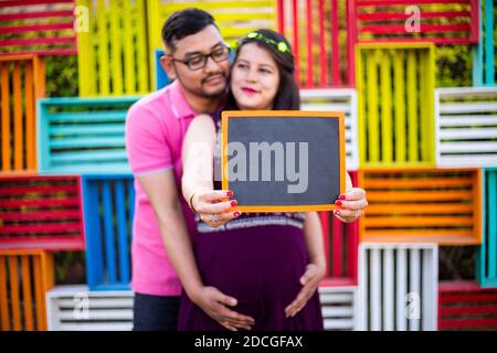 Young pregnant asian indian couple holding writing slate together, pregnancy concept. selective focus on slate. Stock Photo