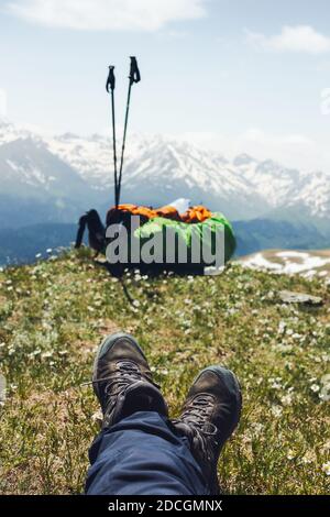First-person photo male climber in sneakers lies on top of hill on green meadow with wildflowers and looks at his backpack and ski poles against a sce Stock Photo