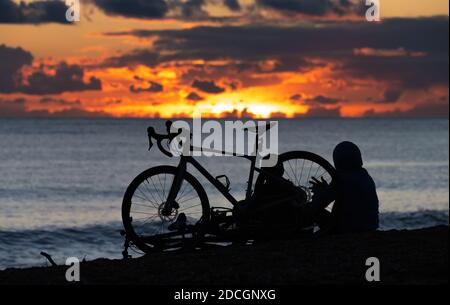 Silhouette of a pair of male cyclists resting on a beach while watching the sun going down. People watching sunset at sea. Stock Photo