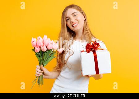 Happy girl in a white T-shirt holding a bouquet of beautiful flowers and a gift box on a yellow background. Stock Photo