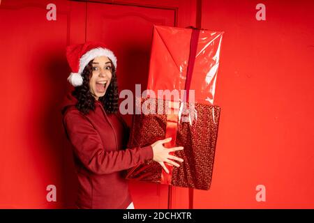 Young woman in Santa Claus hat holds big gifts, standing near red wall. Happy female in Christmas hat with boxes of gift. Concept of holidays, present Stock Photo