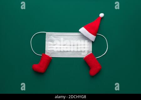 Funny cheerful santa claus from medical mask, red cap and felt boots on green background. Concept for coming christmas and new year 2021 in COVID-19 c Stock Photo