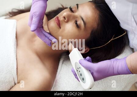 A woman makes an ultrasonic cleaning of the face and skin to a client. Modern equipment. Young pretty woman receiving treatments in beauty salons and lying on the couch. Stock Photo