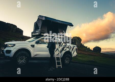 Person admiring a new 'Pathseeker' Auto Solar Hard Shell Roof Top Camping Tent (by Direct4x4 Manufacturing Ltd) on a 4x4 pickup truck with the stunning Cow & Calf Rocks as a landscape backdrop - a growing trend, perhaps in staycations due to COVID-19, Ilkley Moor, England UK Stock Photo