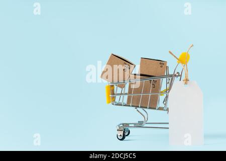 Toy shopping cart with boxes and empty tag on blue background. Copy space for text or design. Sale, discount, shopping and delivery concept. Consumer Stock Photo