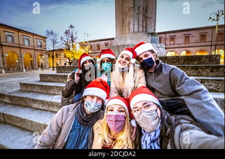 Portrait group of smiling young happy friends wearing face mask and santa hat during Covid pandemic - Multiracial people taking a selfie outdoor havin Stock Photo