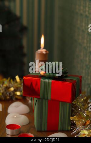 New Year's gifts in red packaging lie on a shelf with candles, in the background a candle is burning and there is a Christmas tree Stock Photo