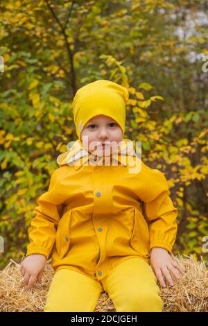 cute beautiful prescholer boy in an orange pants, raincoat, hat, rubber boots sitting on a pile of hay. Cosiness, autumn. Stock Photo