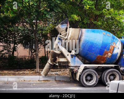 A blue concrete mixer stands on the side of the road near the curb against the backdrop of a house and trees Stock Photo