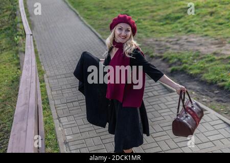 A joyful woman walks up the stairs in burgundy palla and biret, an adult with a charismatic appearance in black clothes, in the fall against a blue Stock Photo