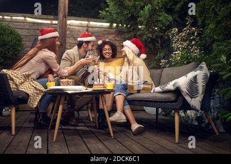 drunk group of young adults drinking white wine, toasting, celebrating new year, wearing santa hats, sitting outdoor in the backyard on the sofa Stock Photo