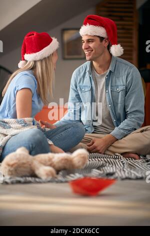 young caucasian couple sitting on a floor at home, looking each other, smiling, wearing santa hats Stock Photo