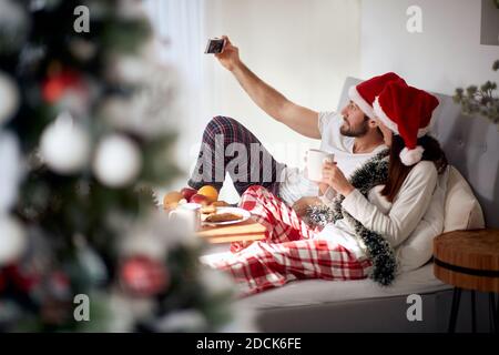 young Caucasian couple having breakfast in bad for Christmas, taking selfie with cell phone, smiling Stock Photo