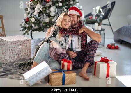 A young ornate couple sitting on the floor at home surrounded by Xmas presents on a beautiful holiday morning. Christmas, relationship, love, together Stock Photo