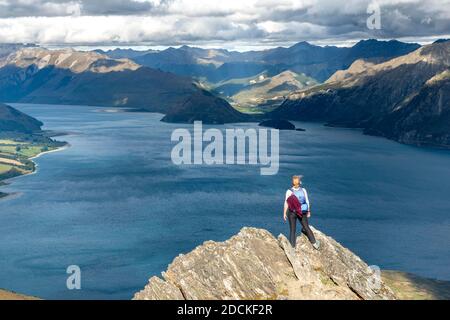 Hiker stands on a rock, view over Lake Hawea, lake and mountain landscape in the evening light, view from Isthmus Peak, Wanaka, Otago, South Island Stock Photo