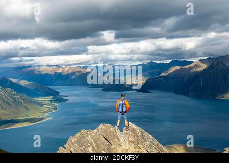 Hiker stands on a rock, view over Lake Hawea, lake and mountain landscape in the evening light, view from Isthmus Peak, Wanaka, Otago, South Island Stock Photo