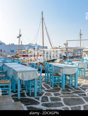 Moored traditional fishing boats and the exterior of a fish tavern with  tables and wooden chairs on the picturesque harbor of Naousa Paros, Greece. Stock Photo