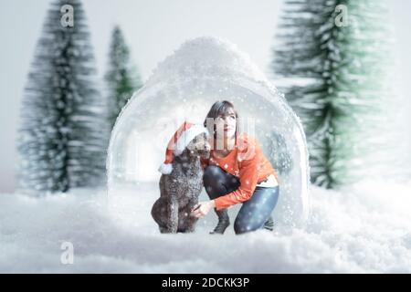 Caucasian woman wearing a christmas sweater and her dog with santa claus hat inside a snow globe. Concept of coronavirus quarantine during christmas s Stock Photo