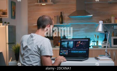 Freelancer editing a video using post production software working from home overtime sitting in the kitchen. Videographer processing audio film montage on professional laptop late at night Stock Photo