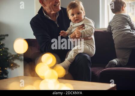 Baby boy with sitting on lap of his grandfather on sofa. Kids with grandparents at home during Christmas. Stock Photo