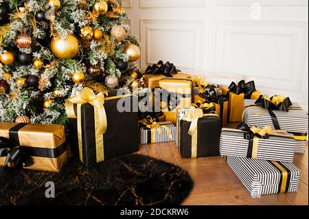 lots of gift boxes in wrapping paper and ribbons in black and gold colors under big fluffy Christmas tree with garland, Christmas card, selective focu Stock Photo