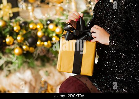 hands of woman in black sequin dress holding big Golden gift with black ribbon, garland and bokeh on Christmas interior on background, selective focus Stock Photo