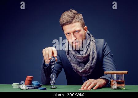 Stylish bearded Man in suit and scarf playing in dark casino, smoking cigar, drink whiskey. Stock Photo