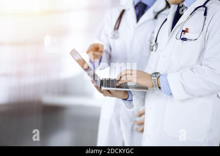 Two unknown doctors with stethoscopes discuss medical exam resoults, while standing at sunny hospital office. Physicians using a laptop for checking Stock Photo