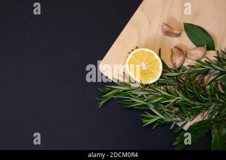 Cutting board of light wood framed with bay leaves, rosemary sprigs, lemon and cloves of garlic, designer template with dark gray background Stock Photo