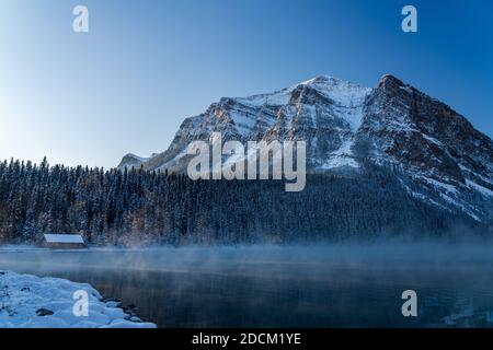 Lake Louise in early winter sunny day morning. Mist floating on turquoise color water surface. Clear blue sky, snow capped Fairview Mountain Stock Photo